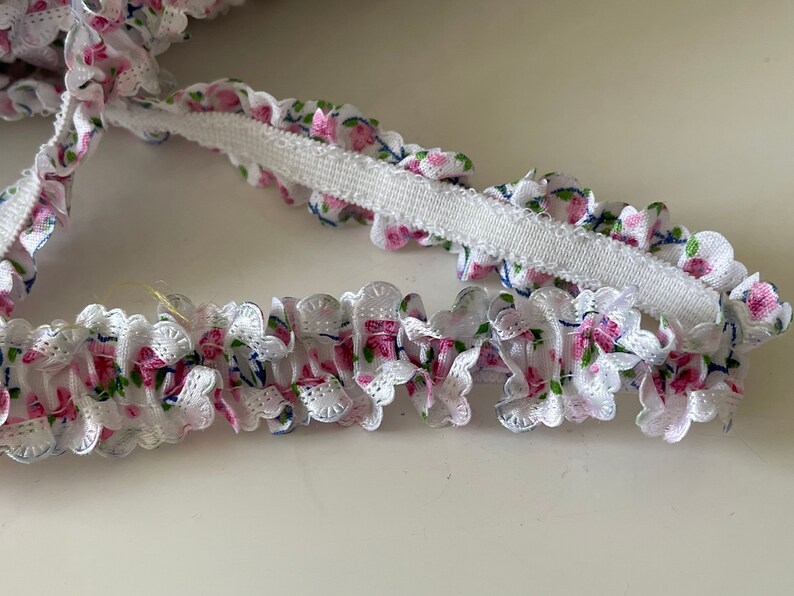 May include: White elastic trim with a pink and blue floral print. The trim has a ruffled edge and is perfect for adding a touch of elegance to any project.