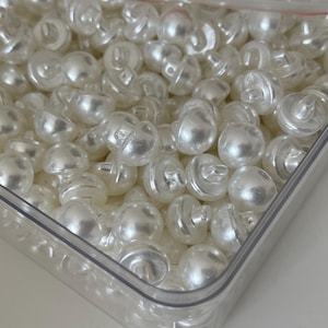 Op de afbeelding: Een doorzichtige plastic container gevuld met witte parelmoerachtige knopen. De knopen hebben een klein gaatje bovenaan om te naaien.