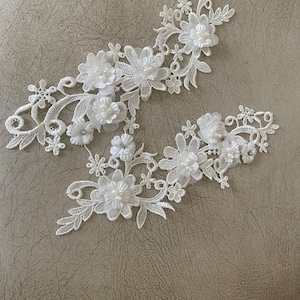 May include: Two white lace floral appliques. Each applique features delicate, three-dimensional flowers with pearl accents and intricate leaf and vine details. The appliques are ideal for embellishing bridal wear or craft projects.