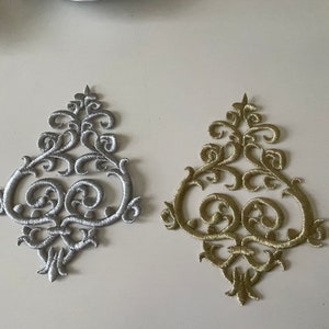 Peut inclure: Deux patchs en tissu décoratifs et ornés dans les couleurs argent et or. Les patchs ont la forme d'un motif stylisé symétrique avec un ovale central et des détails floraux tourbillonnants.