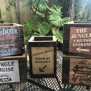 May include: Five wooden boxes with signs on them. The signs are for different attractions at a theme park. The signs read "The Harbour Gallery Come Ashore for Dockside Dining", "Jungle Cruise", "Indiana Jones Stand-By Entrance", "The Jungle Cruise Entrance", and "There it is Ladies & Gentlemen the backside of water".