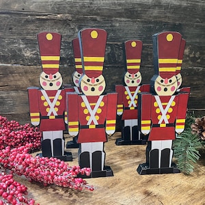 Puede incluir: Cinco adornos de soldados de juguete de madera en rojo, amarillo, blanco y negro. Los soldados están de pie en fila sobre una superficie de madera.