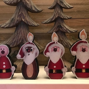 May include: Four wooden Christmas candle holders, each featuring a different character: a snowman, a reindeer, and two Santas. The characters are painted in red, white, and brown with black outlines. The candles are lit and have a yellow flame.