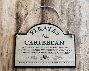 May include: A brown wooden sign with black and green text that reads "Pirates of the Caribbean" and "A thrilling adventure cruise through dark mysterious caverns where "dead men tell no tales" New Orleans Square Disneyland".