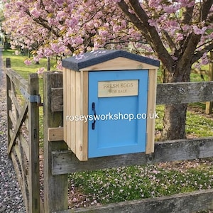 May include: A small wooden egg stand with a blue door and a grey roof, fixed to a wooden fence. The sign on the door reads "FRESH EGGS FOR SALE". The stand is set against a backdrop of a flowering tree.