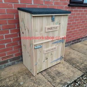 May include: A wooden parcel and wellington boot storage box with a black textured roof. The box has two compartments, one labeled "PARCELS" and the other "BOOTS & WELLIES". The box is made of light-colored wood and has metal hinges and latches.