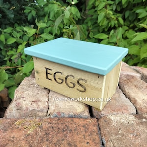 May include: A rectangular wooden storage box with a light blue painted lid and natural wood sides. The front panel features the word EGGS in black lettering and the text roseysworkshop.co.uk printed in smaller black font below it.