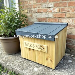 May include: A wooden milk and eggs storage box with a grey slanted roof. The box is made of light-coloured wood with a sign that reads "MILK & EGGS". The website "roseysworkshop.co.uk" is printed on the roof.