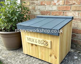 Può includere: Una cassetta di legno per latte e uova con un tetto grigio inclinato. La cassetta è realizzata in legno chiaro con un cartello con la scritta "MILK & EGGS". Il sito web "roseysworkshop.co.uk" è stampato sul tetto.