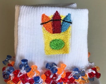 May include: White socks with a colorful embroidered crayon design. The socks have a beaded trim in shades of red, orange, and blue. The embroidered design features a yellow crayon box with red, orange, and blue crayons.