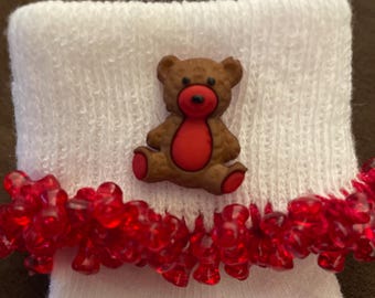 May include: White baby socks with a red beaded trim and a brown teddy bear applique with a red heart.
