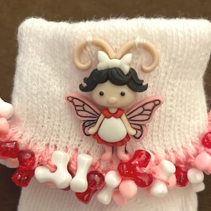 May include: White baby socks with a pink and white fairy button embellishment. The socks are trimmed with red and white beads.