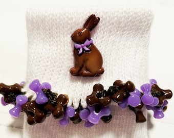 May include: White knitted baby socks with a brown chocolate bunny button and purple bow. The socks are trimmed with brown and purple beads.