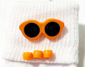 May include: Orange plastic sunglasses with a white fabric background. Three small orange plastic circles are below the sunglasses.