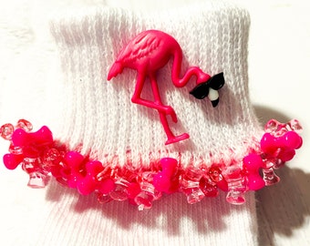 May include: White ankle socks with a pink flamingo charm attached to the top. The flamingo has a black eye and a black beak. The socks have a pink and white ruffle trim.