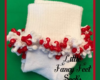 May include: White socks with a decorative red and white trim. The trim is made of small, cross-shaped beads. The text "Little Fancy Feet Socks" is visible in the lower right corner. The background is green with a red border.