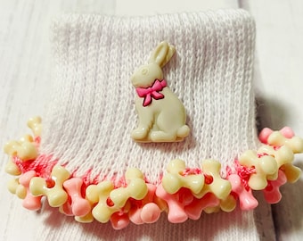 May include: White baby socks with a pink and white bunny button on the top. The socks have a ruffle trim with pink and cream colored beads.