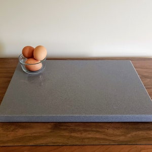 May include: A gray rectangular cutting board with a speckled surface sits on a wooden table. A small glass bowl with three brown eggs is in front of the board.