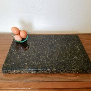May include: A dark green granite cutting board with a slightly rough surface. The board is sitting on a wooden table with a small glass bowl of eggs in the background.