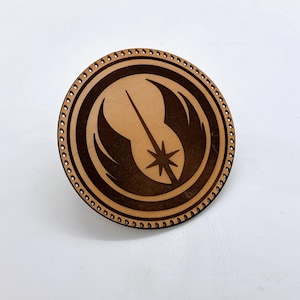 May include: A round, wooden pin with a laser-etched design. The pin features a central emblem with a star and a stylized wing design, surrounded by a circular border with small, evenly spaced holes. The wood has a light brown color with darker brown etched details.