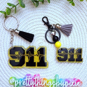 May include: Two keychains featuring the number 911 in black with a yellow glitter background and a heartbeat line graphic. One keychain has a silver ring and black tassel, while the other has a black clip, beads, and a gray tassel. A sign reads @prettythingsbypixie.