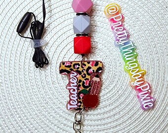 May include: A teacher lanyard with a leopard print charm that says "Teacher" with a red apple and a pencil. The lanyard has a gray, pink, and red silicone beads and a black breakaway clasp.