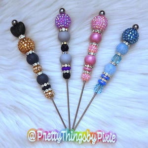 May include: Five colorful beaded stirring sticks with metal tips. The beads are various colors, including pink, blue, purple, and gold. The sticks are all the same length and have a silver metal tip. The sticks are arranged in a row on a white fluffy surface. The text "@PrettyThingsbyPixie" is visible at the bottom of the image.