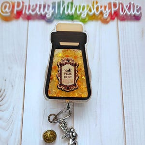 May include: A gold glitter filled badge reel with a clear plastic jar charm that says "Pixie Dust" and a silver fairy charm.