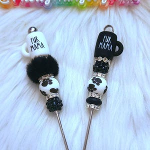 May include: Two black and white coffee mug charm keychains with the text "Fur Mama" on the mugs. The charms are made with black and white beads, rhinestones, and a black fur pom-pom.