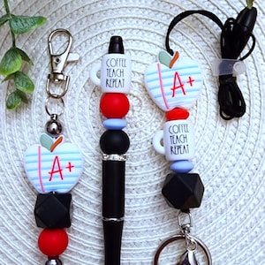 May include: A black pen with a silver accent and a red, blue, and black beaded keychain with a white apple charm that says "A+" and a white coffee mug charm that says "Coffee Teach Repeat".