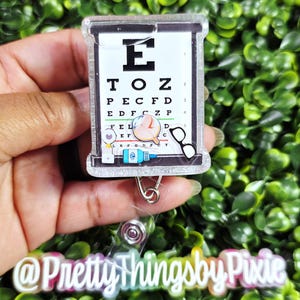 May include: A rectangular badge reel featuring a white eye chart with black letters and numbers. The design includes small colorful icons of an eye, a magnifying glass, a bottle of eye drops, and black glasses. The text @PrettyThingsbyPixie is visible.