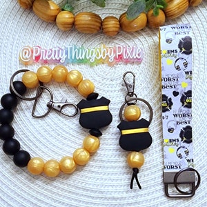 May include: A black and gold beaded bracelet with a black and yellow badge charm. A matching keychain with a black and yellow badge charm is also pictured. The keychain has a black and white ribbon with the text "Tell me your worst, I'll send the best" and "EMS Dispatcher" repeated on it.