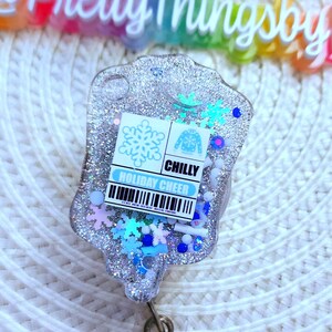 May include: A silver glitter resin badge holder with a blue snowflake and a blue sweater design. The badge holder has a blue and white label that reads "CHILLY HOLIDAY CHEER".