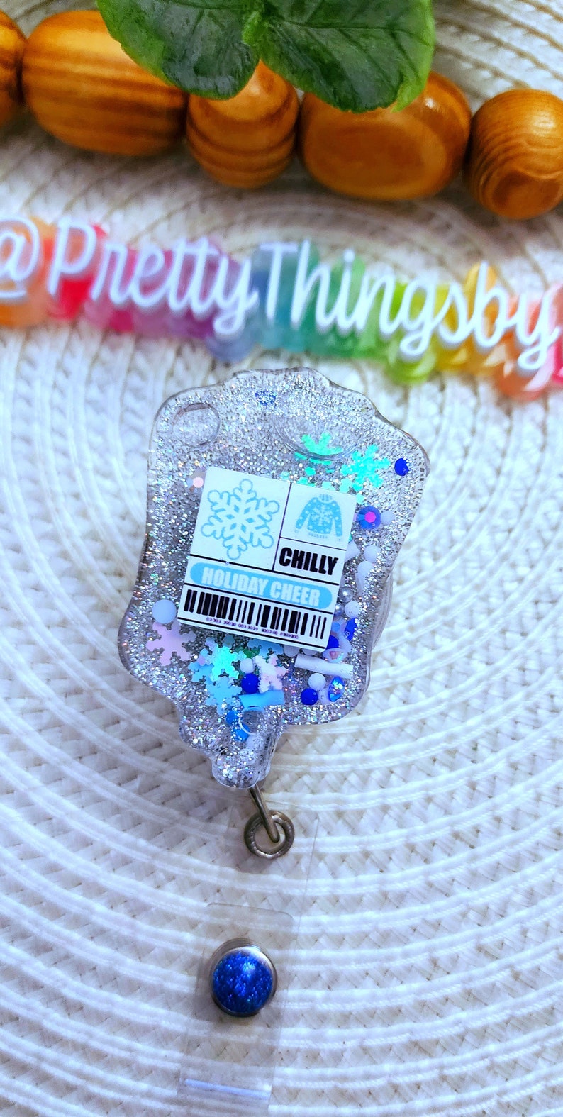 May include: A silver glitter resin badge holder with a blue snowflake and a blue sweater design. The badge holder has a blue and white label that reads "CHILLY HOLIDAY CHEER".