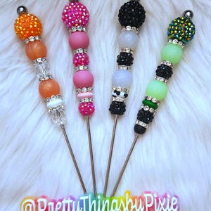 May include: Four colorful, jeweled, and beaded stirring sticks with metal tips. The sticks are arranged in a row on a white fluffy surface. The text "@PrettyThingsbyPixie" is visible at the bottom of the image.