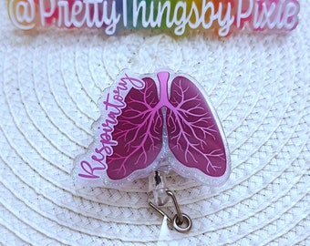 May include: A badge reel featuring a translucent acrylic topper shaped like human lungs in shades of dark pink and purple with the word Respiratory written in cursive script. The topper has a glittery border and a silver metal clip with a clear strap.