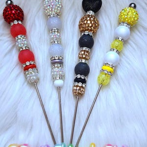 May include: Four decorative, beaded, metal stirring sticks. The sticks are topped with a crown, a heart, a yellow bead, and a red bead. The beads are various colors and sizes. The sticks are on a white fuzzy surface. The text "@PrettyThingsbyPixie" is at the bottom of the image.