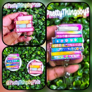May include: A badge reel featuring a stack of colorful books with floral and patterned spines. The books are rendered in shades of pink, blue, yellow, and teal with glitter accents. A metal clip is attached to the bottom of the acrylic book stack.