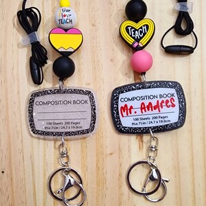 May include: Two teacher themed keychains with black lanyards, colorful beads, and a composition book charm. The charms feature the words "Live Love Teach" and "Teach". The composition book charms have glitter and the text "100 Sheets 200 Pages 9 1/2 x 7 1/2 in / 24.7 x 19.0 cm". The keychains are attached to silver key rings.