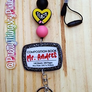 May include: A lanyard featuring a black cord, a black bead, a heart-shaped bead with the text TEACH, a pink bead, and a rectangular pendant resembling a composition book labeled Mr. Andres with 100 Sheets 200 Pages 9.75 x 7.5 in / 24.7 x 19.0 cm.