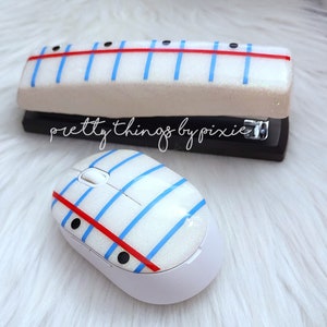May include: A white stapler and a white computer mouse with a blue and red striped design. The design resembles a sheet of lined paper. The text "pretty things by pixie" is visible on the stapler.