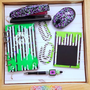 May include: A black and white striped notebook with green slime dripping down the cover. The notebook says "Strange and Unusual". There are also black and white striped paper clips, a black stapler with green and purple splatter paint, a black computer mouse with green and purple splatter paint, and a black pen with a green and purple beetle charm.