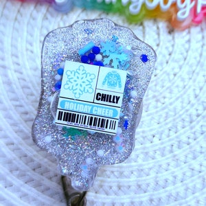 May include: A silver glitter resin badge holder with a blue and white holiday cheer design. The badge holder has a snowflake and a Christmas sweater with the word "CHILLY" on it. The badge holder is attached to a clear plastic badge reel with a blue glitter button.