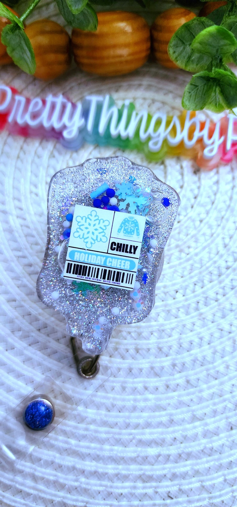 May include: A silver glitter resin badge holder with a blue and white holiday cheer design. The badge holder has a snowflake and a Christmas sweater with the word "CHILLY" on it. The badge holder is attached to a clear plastic badge reel with a blue glitter button.