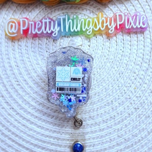 May include: A silver glitter badge holder with a blue snowflake and sweater design. The badge holder has a clear retractable reel with a blue glitter button. The text "CHILLY HOLIDAY CHEER" is printed on the badge holder.