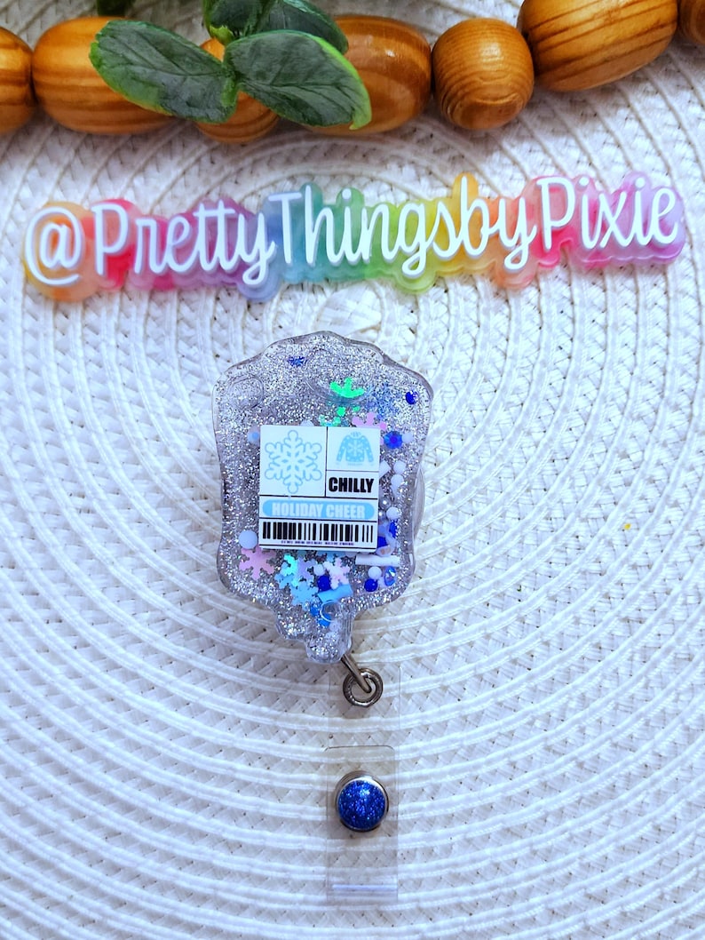 May include: A silver glitter badge holder with a blue snowflake and sweater design. The badge holder has a clear retractable reel with a blue glitter button. The text "CHILLY HOLIDAY CHEER" is printed on the badge holder.