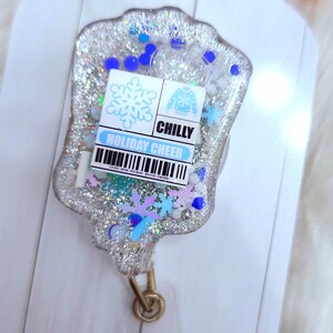 May include: A silver glitter resin keychain with a blue snowflake, a blue sweater, and the text "CHILLY HOLIDAY CHEER".