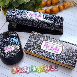 May include: A set of school supplies featuring a black and white speckled design. The set includes a stapler, a mouse, and a composition book. Each item has a label that reads "Composition Book" and "Ms. Betty".