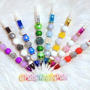 May include: Nine colorful beaded pens with clear plastic tops. The beads are various colors, including white, blue, green, pink, and purple. The pens are arranged in a semi-circle on a white fuzzy surface. The text "@PrettyThingsbyPixie" is visible at the bottom of the image.