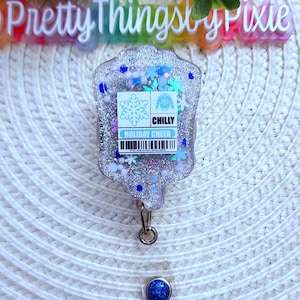 May include: A silver glitter badge holder with a blue snowflake and a white sweater with the text "CHILLY HOLIDAY CHEER". The badge holder has a blue glitter retractable badge reel.